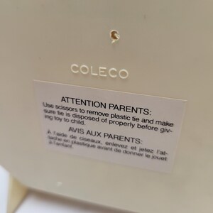 May include: Cream-colored plastic toy with the brand name "COLECO" printed on the surface. A white sticker with text in English and French provides safety instructions for parents.