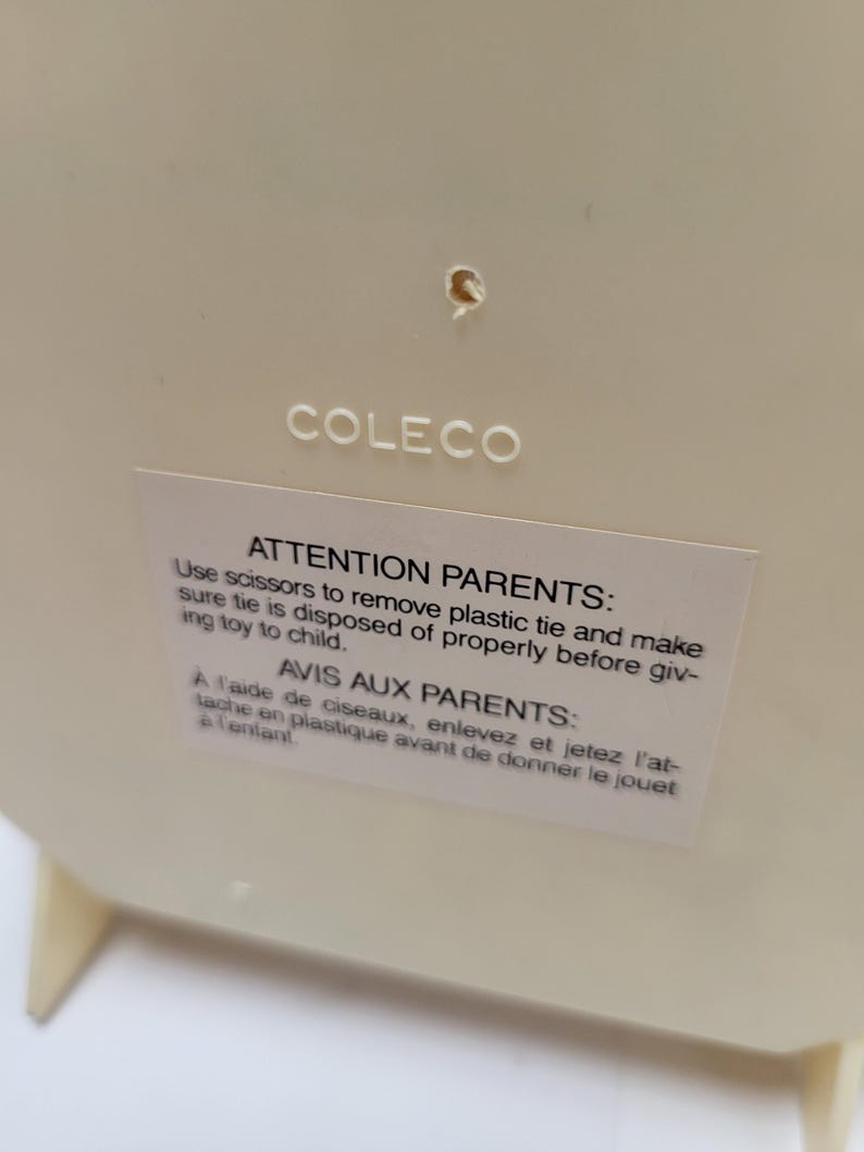 May include: Cream-colored plastic toy with the brand name "COLECO" printed on the surface. A white sticker with text in English and French provides safety instructions for parents.