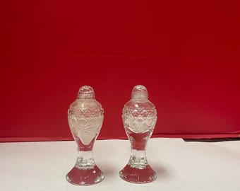 May include: Two clear glass salt and pepper shakers with a decorative design. Each shaker has a bulbous body, a slender neck, and a flared base. The shakers are topped with a clear, rounded stopper. The background is a solid red.