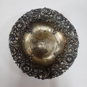 Puede incluir: Cuenco antiguo plateado con un borde ornamentado de flores y volutas. El interior del cuenco tiene un monograma y un acabado ligeramente deslustrado. Una pieza decorativa para servir o exhibir.