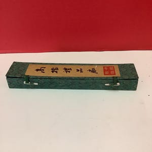 K&ouml;nnte beinhalten: Eine gr&uuml;ne Box mit einem braunen Etikett, auf dem in chinesischen Schriftzeichen "Chinesischer Kalligraphie-Pinsel" steht. Die Box hat zwei wei&szlig;e Verschl&uuml;sse an beiden Seiten.