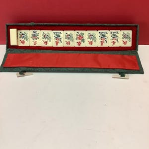 K&ouml;nnte beinhalten: Ein gr&uuml;nes Stoffetui mit roter Innenausstattung enth&auml;lt einen Satz von zehn wei&szlig;en Mahjong-Steinen mit chinesischen Schriftzeichen und Blumendesigns.