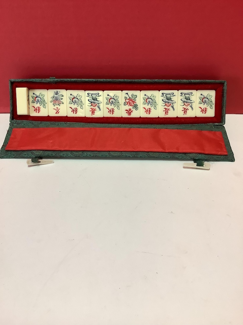 K&ouml;nnte beinhalten: Ein gr&uuml;nes Stoffetui mit roter Innenausstattung enth&auml;lt einen Satz von zehn wei&szlig;en Mahjong-Steinen mit chinesischen Schriftzeichen und Blumendesigns.