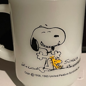 May include: White ceramic mug with a black and white illustration of Snoopy and Woodstock. The mug has the text "Copr. © 1958, 1965 United Feature Syndicate, Inc." on the bottom.
