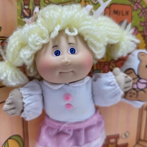 May include: A vintage doll with blonde yarn hair styled in pigtails. The doll has blue eyes, rosy cheeks, and is wearing a white top with pink buttons and a pink skirt with white polka dots and lace trim. The doll is inside a cardboard box.