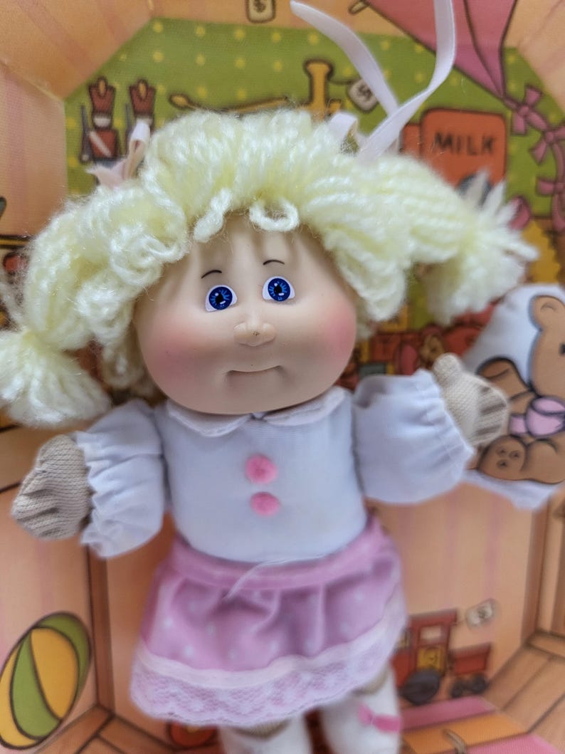 May include: A vintage doll with blonde yarn hair styled in pigtails. The doll has blue eyes, rosy cheeks, and is wearing a white top with pink buttons and a pink skirt with white polka dots and lace trim. The doll is inside a cardboard box.