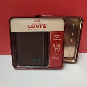 Op de afbeelding: Een bruine leren Levi's portemonnee in een rode en gouden blikken doos. De portemonnee zit in een doorzichtige plastic hoes met een kartonnen achterkant. De doos heeft het Levi's logo en de tekst "America's Original Jeans Since 1973". RFID-bescherming.