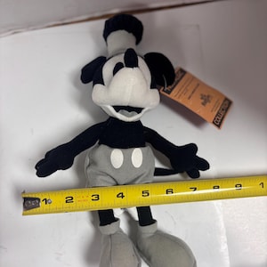 May include: A black, white, and gray Mickey Mouse plush toy. The plush has a black hat, ears, and arms, a white face, and gray shorts and shoes. The toy is approximately 6 inches tall. A yellow measuring tape is next to the toy.