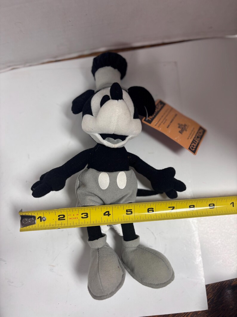 May include: A black, white, and gray Mickey Mouse plush toy. The plush has a black hat, ears, and arms, a white face, and gray shorts and shoes. The toy is approximately 6 inches tall. A yellow measuring tape is next to the toy.