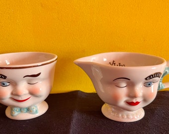 Könnte beinhalten: Zwei weiße Keramikbecher mit Gesichtern. Ein Becher hat ein zwinkerndes Gesicht mit einem braunen Schnurrbart und dem Text "BAILEYS" an der Seite. Der andere Becher hat ein zwinkerndes Gesicht mit roten Lippen und dem Text "1/2 pint" im Inneren.