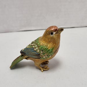 Op de afbeelding: Een decoratief vogelbeeldje met een gouden basis. Het heeft een bruine kop, groene en gouden vleugels en een blauwe rug. De vleugels zijn versierd met kleine, heldere kristallen. De snavel en poten van de vogel zijn goudkleurig.