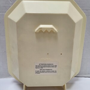 May include: Cream-colored plastic toy with a geometric shape and a built-in hanger. The back of the toy has a label with text in English and French, including a warning for parents. The toy has two small legs for standing.