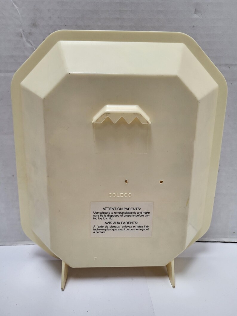 May include: Cream-colored plastic toy with a geometric shape and a built-in hanger. The back of the toy has a label with text in English and French, including a warning for parents. The toy has two small legs for standing.