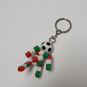 May include: A keychain featuring a figure constructed from small red, white, and green cubic blocks. The figure has a round white head with black pentagonal soccer ball patterns. The entire assembly is attached to a circular metal split ring.