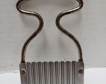 Op de afbeelding: Een vintage metalen crinkle cutter met een golvend mes en een gebogen handvat. Het mes is rechthoekig met verticale ribbels en het handvat heeft een lusvorm. Het metaal heeft een aangetaste, verouderde uitstraling.
