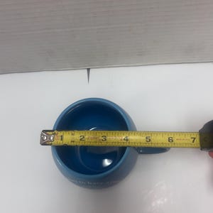 May include: A blue ceramic mug with a handle, measuring approximately 4 inches in diameter. The mug has a rounded shape and a smooth, glossy finish. The text on the mug is partially visible. A yellow measuring tape is placed across the mug.