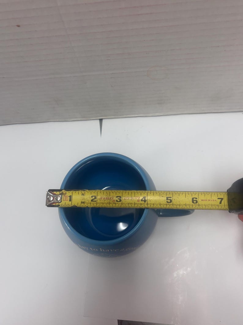 May include: A blue ceramic mug with a handle, measuring approximately 4 inches in diameter. The mug has a rounded shape and a smooth, glossy finish. The text on the mug is partially visible. A yellow measuring tape is placed across the mug.