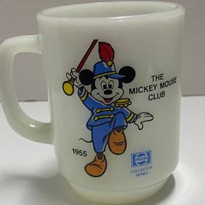 May include: White ceramic mug with a cartoon image of Mickey Mouse in a blue and gold uniform, holding a red and gold baton. The mug has the text "The Mickey Mouse Club" and "1955" printed on it. The bottom of the mug has a Pepsi logo and the text "Collector Series".