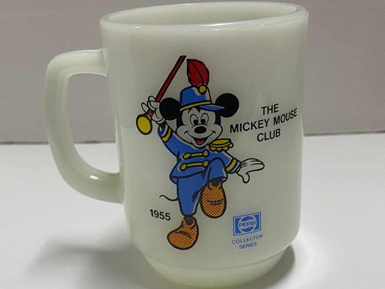 May include: White ceramic mug with a cartoon image of Mickey Mouse in a blue and gold uniform, holding a red and gold baton. The mug has the text "The Mickey Mouse Club" and "1955" printed on it. The bottom of the mug has a Pepsi logo and the text "Collector Series".