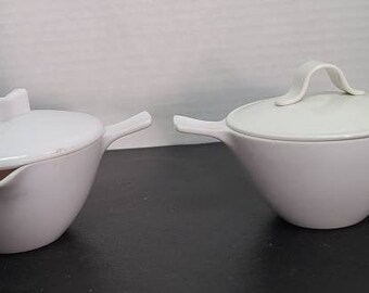 May include: A white ceramic creamer and sugar bowl set with lids and handles. The creamer has a spout for pouring.