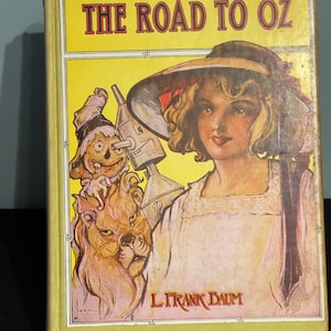 Może przedstawiać: Okładka starej książki zatytułowanej "The Road to Oz" autorstwa L. Franka Bauma. Na okładce przedstawiono młodą dziewczynę w słomkowym kapeluszu, lwa, straszak i blaszanego człowieka.