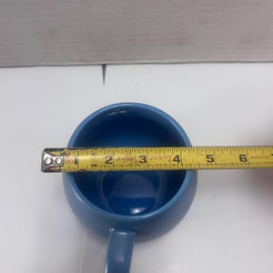 May include: A blue ceramic mug with a handle, measuring approximately 4 inches in diameter. The interior is a darker shade of blue than the exterior. A yellow measuring tape is placed across the mug's opening.