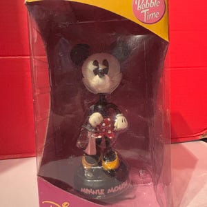 May include: A Minnie Mouse bobblehead figure in a clear plastic display case. The figure is wearing a red and white polka dot dress and yellow shoes. The packaging reads "Collector's Edition" and "Bobble Time."