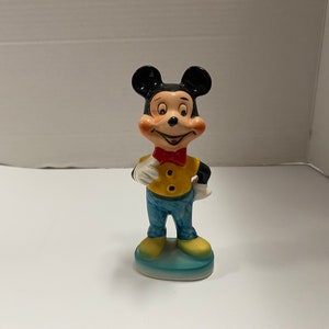 May include: A ceramic figurine of Mickey Mouse wearing a yellow shirt, blue pants, and a red bow tie. He is standing on a blue base.