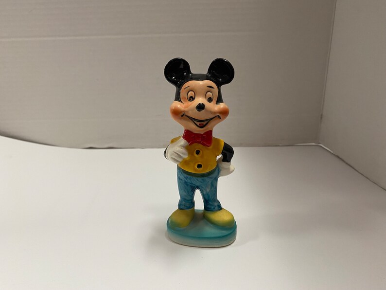 May include: A ceramic figurine of Mickey Mouse wearing a yellow shirt, blue pants, and a red bow tie. He is standing on a blue base.