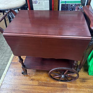 May include: A dark cherry-colored wooden tea cart with a rectangular top and a lower shelf. The cart features a decorative spoked wheel and a handle. The cart is designed for serving and displaying items.