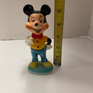 May include: A ceramic figurine of Mickey Mouse wearing a yellow shirt, blue pants, and a red bow tie. He is standing on a blue base and is approximately 5 inches tall.