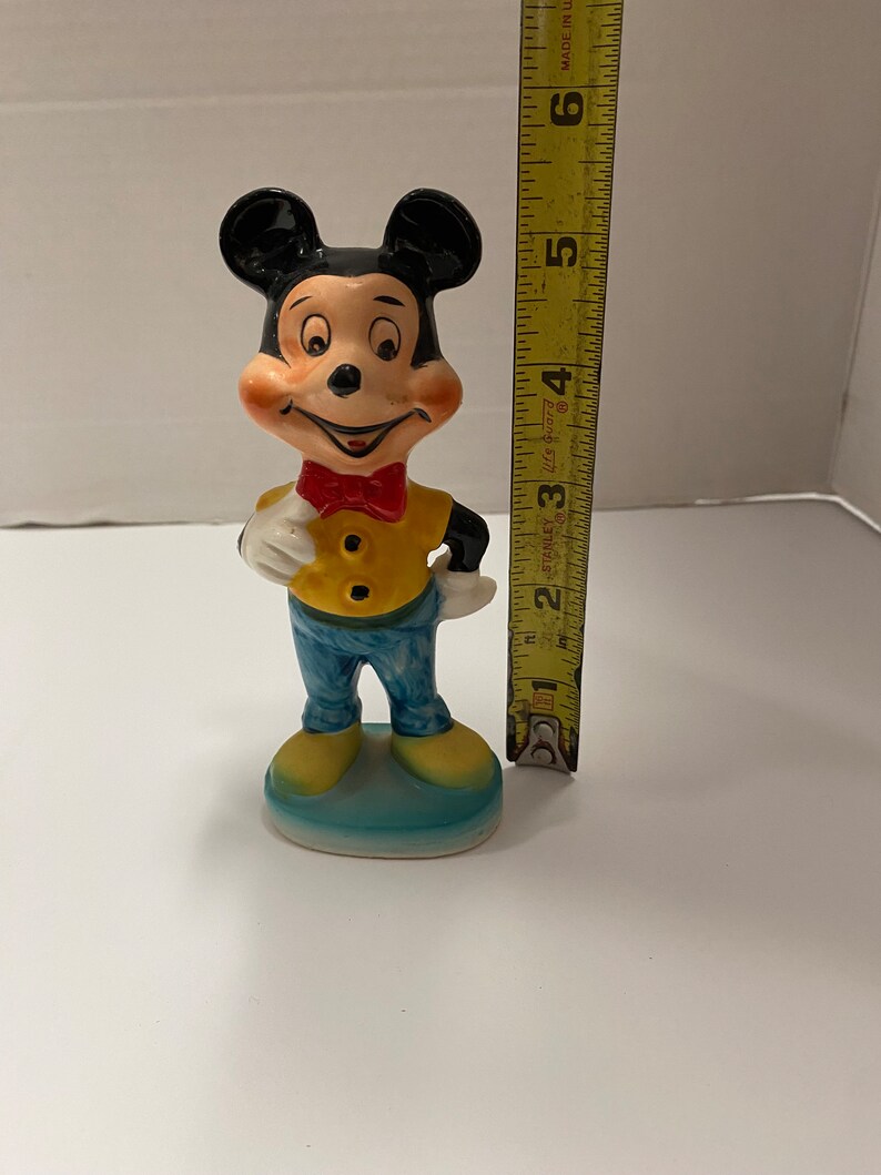 May include: A ceramic figurine of Mickey Mouse wearing a yellow shirt, blue pants, and a red bow tie. He is standing on a blue base and is approximately 5 inches tall.