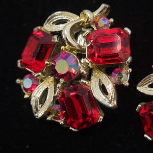 May include: Close-up of a pair of vintage clip-on earrings. The earrings feature large, rectangular, and round red gemstones set in a gold-tone metal. The design includes gold-tone leaves and swirls, with smaller iridescent stones.