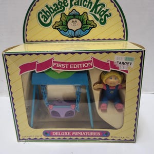May include: A vintage Cabbage Patch Kids Deluxe Miniatures set, featuring a doll and a swing set, in its original packaging. The box is yellow with the words "First Edition" and "Deluxe Miniatures". The doll is wearing blue overalls.
