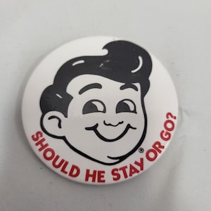 May include: A white button with a cartoon illustration of a smiling boy with black hair. The text "SHOULD HE STAY OR GO?" is printed in red around the edge of the button.