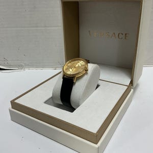 Pu&ograve; includere: Un orologio da polso color oro con cinturino in pelle nera, caratterizzato da un quadrante rotondo con motivo a raggiera testurizzato e lancette dorate. L'orologio &egrave; esposto su un supporto imbottito bianco in una scatola color crema con VERSACE.