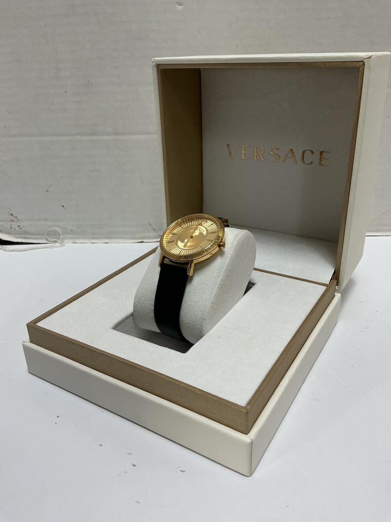 Pu&ograve; includere: Un orologio da polso color oro con cinturino in pelle nera, caratterizzato da un quadrante rotondo con motivo a raggiera testurizzato e lancette dorate. L'orologio &egrave; esposto su un supporto imbottito bianco in una scatola color crema con VERSACE.