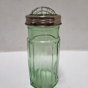 May include: A vintage green glass flower frog with a metal lid and wire grid. The jar has a ribbed design and a hexagonal base. The lid is silver-colored. This item is suitable for floral arrangements.