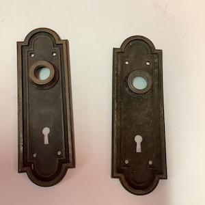 May include: Two antique brass door plates with a keyhole and a round hole for a knob. The plates have a decorative border and are in good condition.