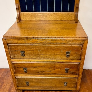 May include: A vintage wooden dresser with three drawers and a top section featuring dark blue tiles. The dresser has a warm, golden-brown finish, ornate drawer pulls, and a classic design. The piece is likely made of oak or a similar hardwood.