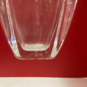 May include: Clear glass vase with a geometric shape and a slightly iridescent finish. The vase has a square base and a rectangular body.