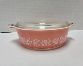 May include: A round, coral-colored glass casserole dish with two side handles and a clear glass lid. The exterior of the dish features a repeating white botanical pattern of leaves and berries encircling the entire base of the vessel.
