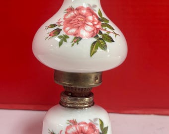 May include: White oil lamp with a floral rose design in pink and green. The lamp has a bulbous top and base, connected by a metal fixture. The lamp is a decorative item.