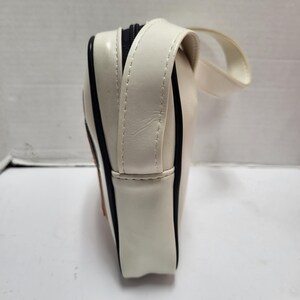 May include: Cream-colored rectangular bag with a black zipper and a small handle. The bag has black trim along the edges and a stitched seam detail. The bag is made of a glossy material.