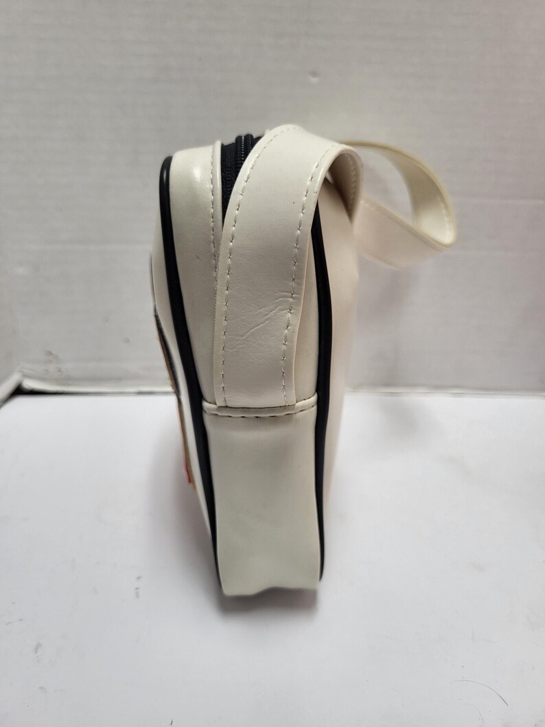 May include: Cream-colored rectangular bag with a black zipper and a small handle. The bag has black trim along the edges and a stitched seam detail. The bag is made of a glossy material.
