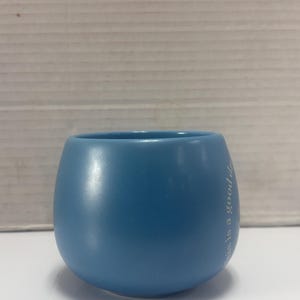 May include: A light blue ceramic tumbler with a rounded base and a wide opening. The text "this is a good day" is printed vertically in white on the side of the tumbler. The tumbler is sitting on a white surface.