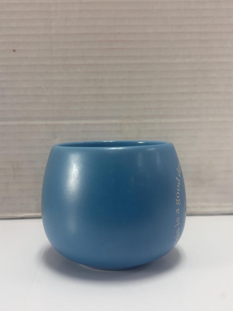 May include: A light blue ceramic tumbler with a rounded base and a wide opening. The text "this is a good day" is printed vertically in white on the side of the tumbler. The tumbler is sitting on a white surface.