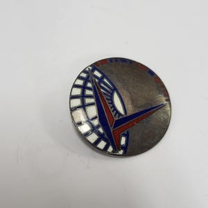 May include: A circular metal pin featuring a stylized design with a grid pattern in white and blue. A prominent red and blue V-shaped element overlays the grid. The remaining portion of the circle has a dark, tarnished metallic finish with small red and blue rectangular accents along the outer edge.