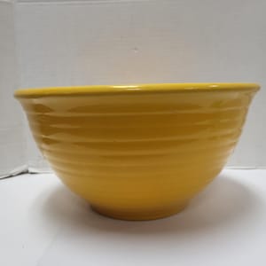 Puede incluir: Un cuenco para mezclar de cerámica amarillo brillante. El cuenco tiene forma redondeada con un borde ancho y una serie de estrías horizontales en el exterior. Está sobre una superficie blanca.