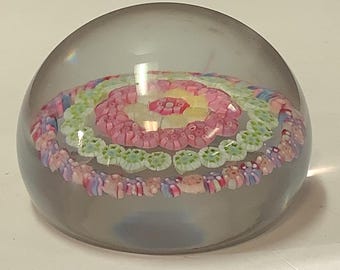 May include: A clear glass paperweight with a multi-colored swirl design. The swirl is made up of pink, yellow, green, and blue glass.